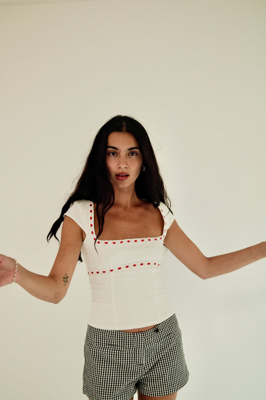 White linen fitted top with square neckline and layered broderie anglaise trimming with red ribbon detail, styled with grey check shorts, model standing against a warm neutral background.