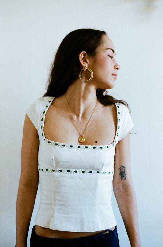 Three-quarter side view of white linen Elissa Top with square neckline and broderie anglaise trim with green ribbon detail, model against a neutral background.