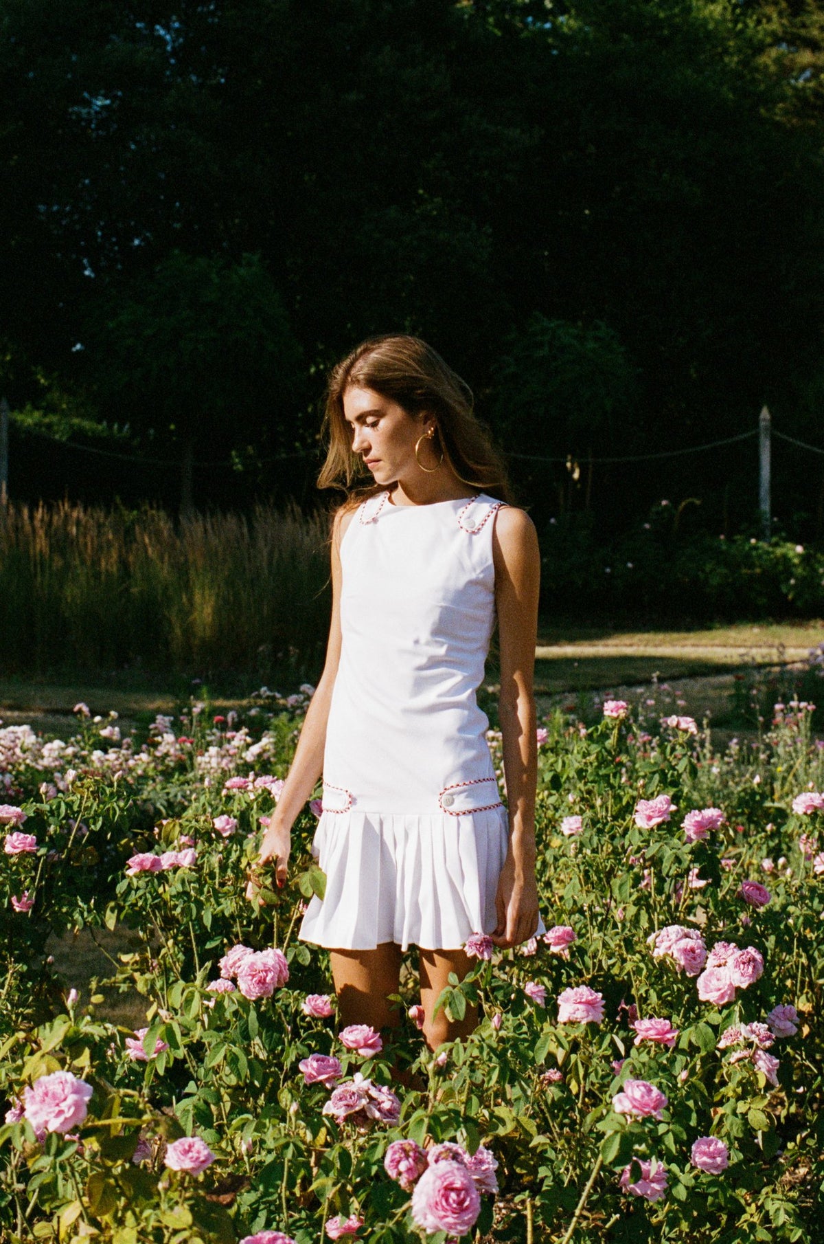 A woman wearing a sleeveless white pleated dress stands in a sunlit garden filled with pink flowers, with dark green trees in the background.