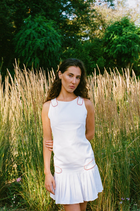A woman wearing a sleeveless white mini dress with red embroidered details stands in a field of tall dry grass with green trees in the background.