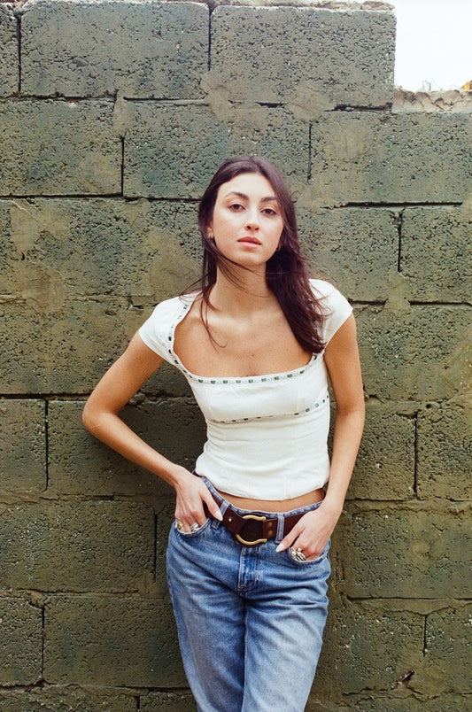 White linen fitted top with square neckline and layered broderie anglaise trimming with green ribbon detail, styled with blue jeans, model standing against a stone wall.