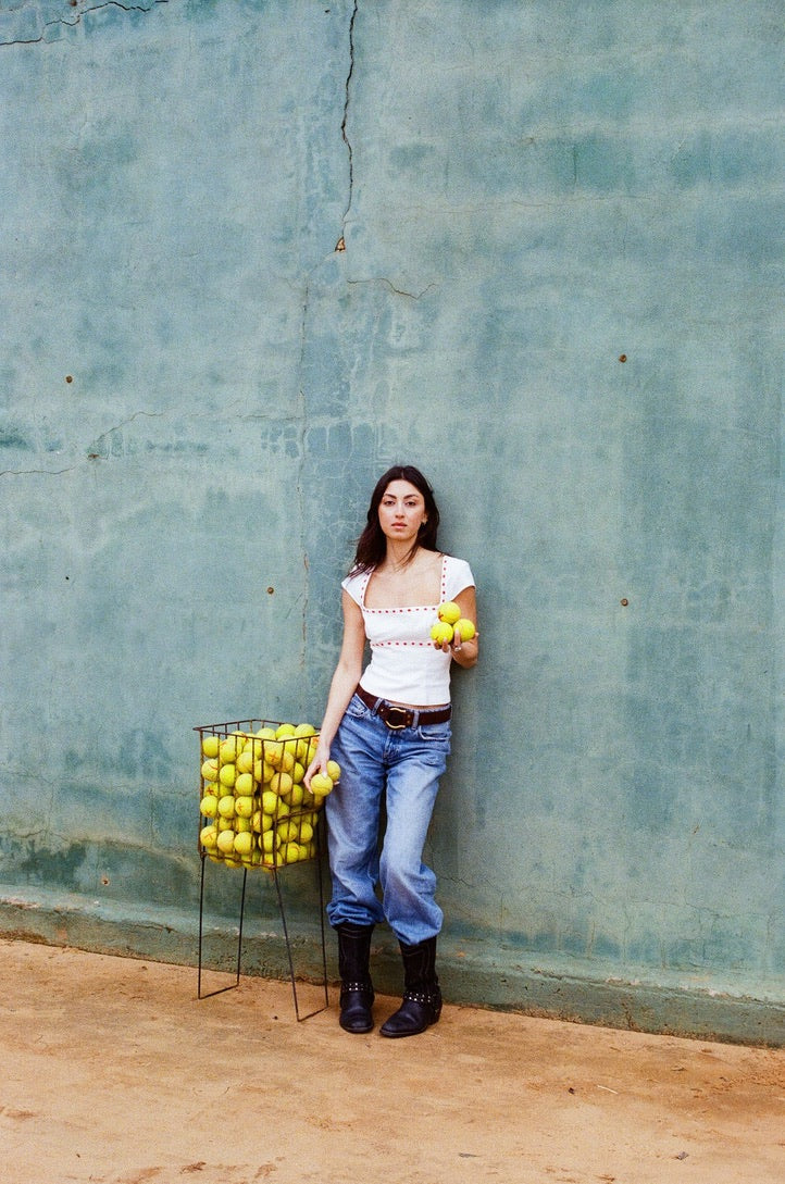 Full-length view of white linen Norma Top with square neckline and red ribbon broderie anglaise trim, styled with blue jeans, model standing against a blue wall.
