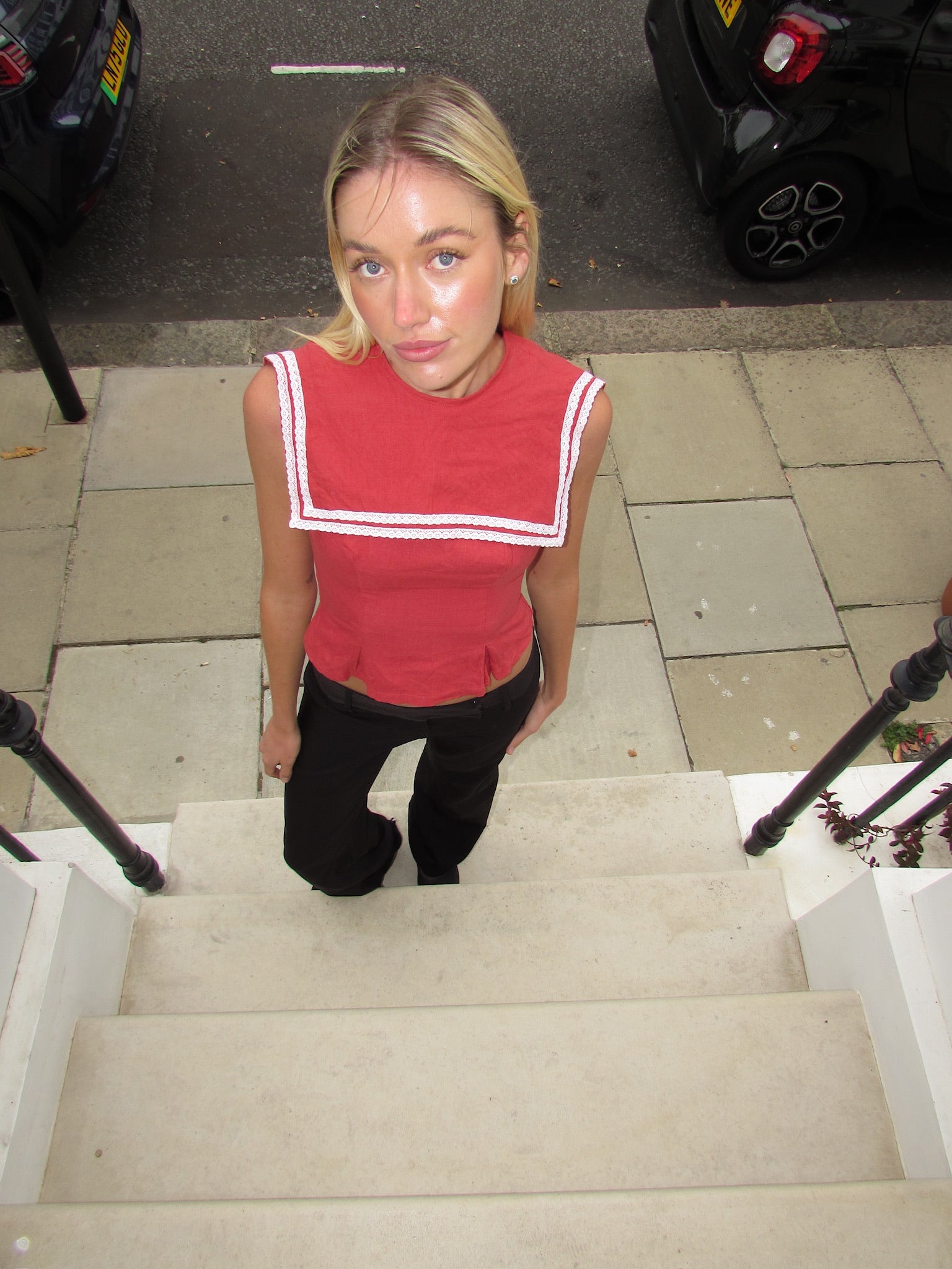 Red linen crop top with white square sailor collar and white stripe trim, worn with black trousers, shot from above on concrete stairs.
