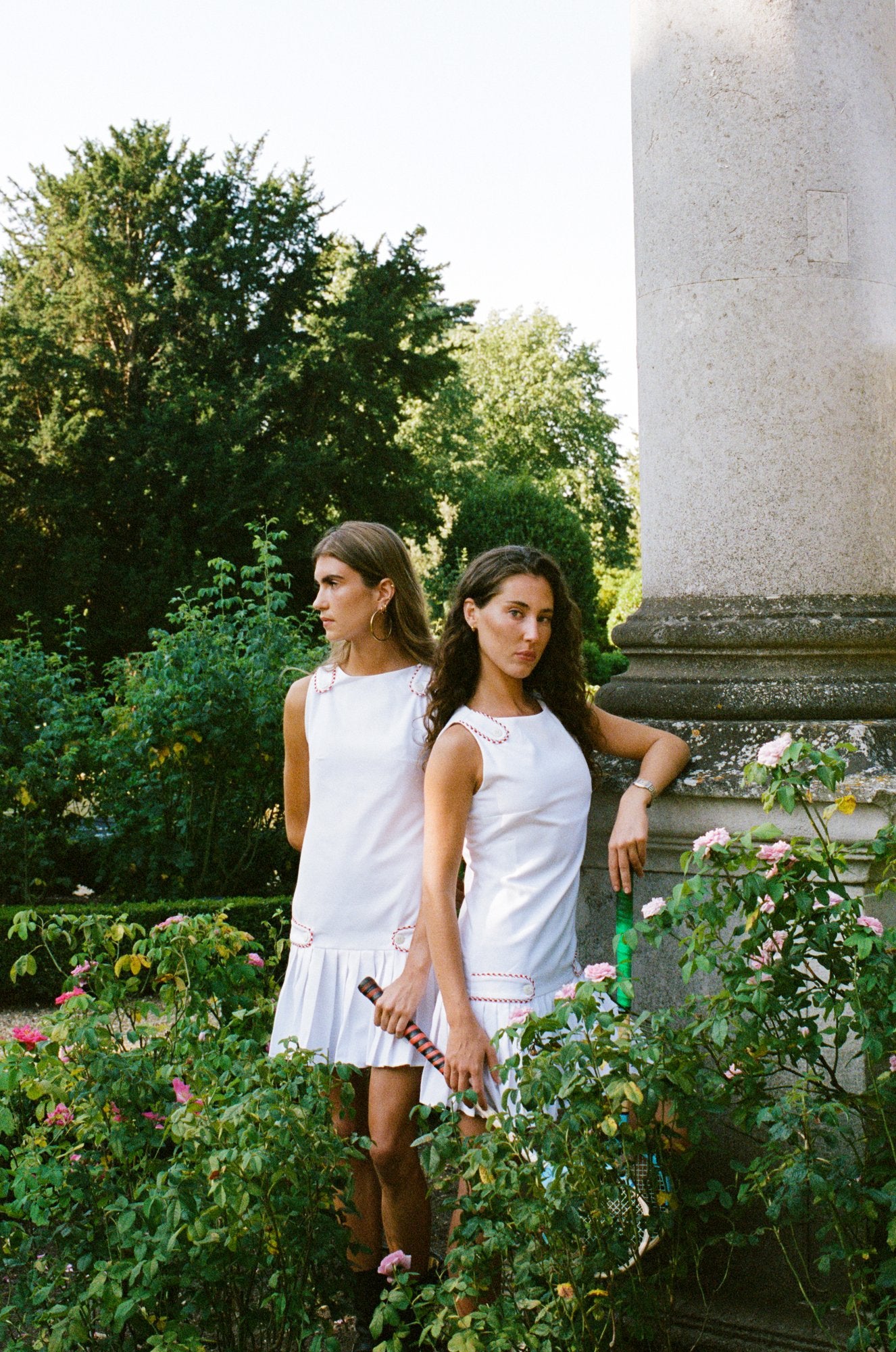Two women wearing white tennis dresses beside a stone column in a garden, one holding a wooden racket.