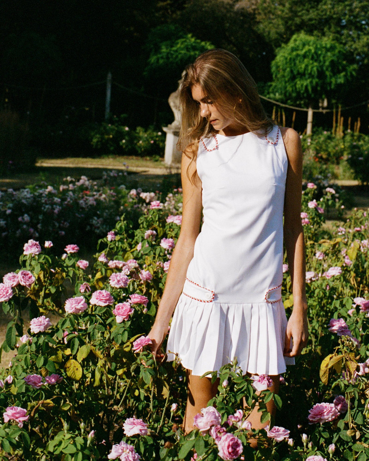 A woman wearing a white sleeveless mini dress with pink embroidered details stands in a sunlit rose garden surrounded by pink flowers.
