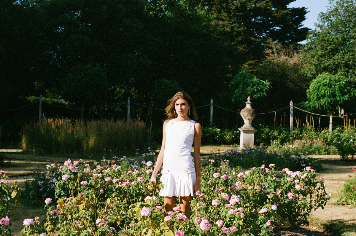 A woman wearing a white sleeveless dress stands in a sunlit garden bed filled with pink flowers, with trees, a rope fence, and a stone urn in the background.