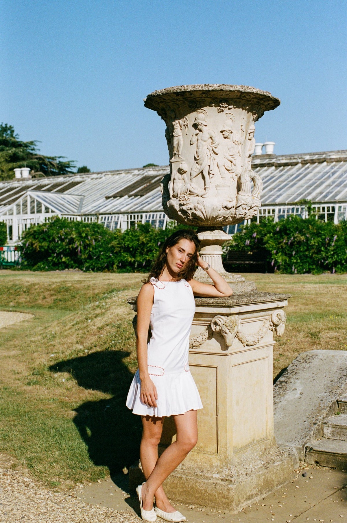A woman in a white sleeveless mini dress stands outdoors beside a large carved stone urn on a pedestal, with grass, shrubs, and a greenhouse in the background under a blue sky.
