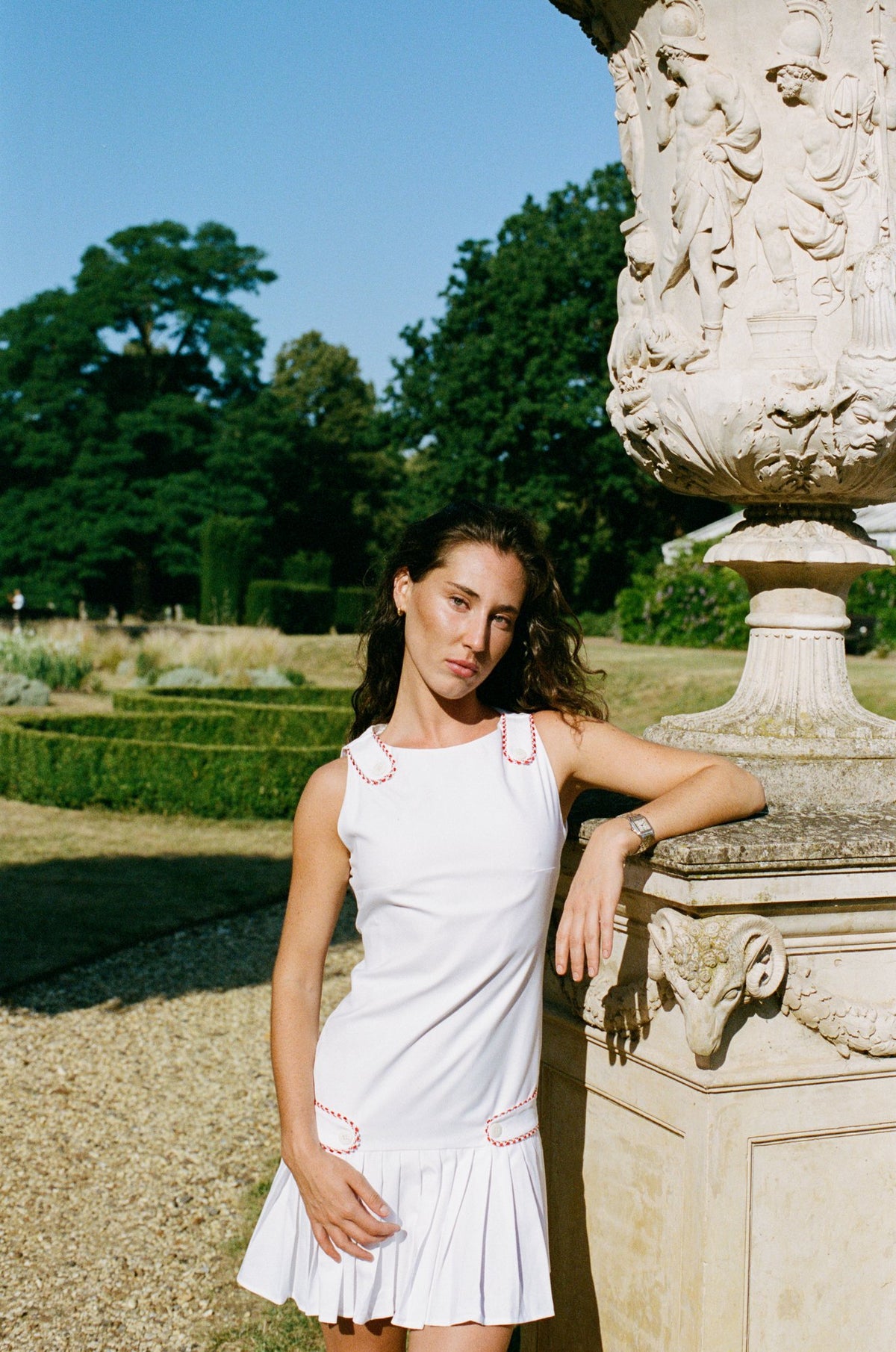 A woman wearing a white sleeveless pleated dress with red embroidered accents leans against a stone garden urn in a sunny park setting.